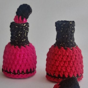 May include: Two crocheted nail polish bottle shaped ornaments. One is pink with black trim and a black top. The other is red with black trim and a black top. Both have gold glitter accents.