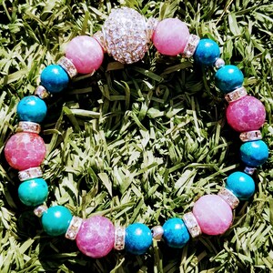 May include: A beaded bracelet featuring alternating round pink and turquoise beads, interspersed with small silver-toned spacers encrusted with clear crystals, and a single larger spherical focal bead covered in sparkling clear rhinestones.