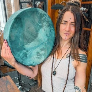 May include: A large, circular, hand-held drum with a textured, deep teal-coloured drumhead. The drum features a solid, matching teal-coloured frame and a smooth, matte finish across the surface, presenting a consistent, monochromatic appearance.