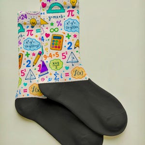 May include: A pair of crew socks featuring a white background with a colorful math-themed print including equations, calculators, light bulbs, and geometric shapes. The heel, toe, and foot section of the socks are solid black in color.
