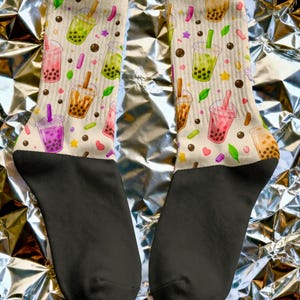 May include: A pair of crew socks with a cream-colored background featuring a repeating pattern of bubble tea cups in various colors including green, pink, purple, and brown, interspersed with small colorful shapes like stars, hearts, and leaves.