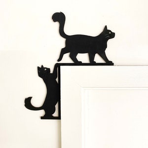 May include: Two black metal cat silhouettes mounted on a white door frame. The cats are positioned as if they are climbing on the door frame.