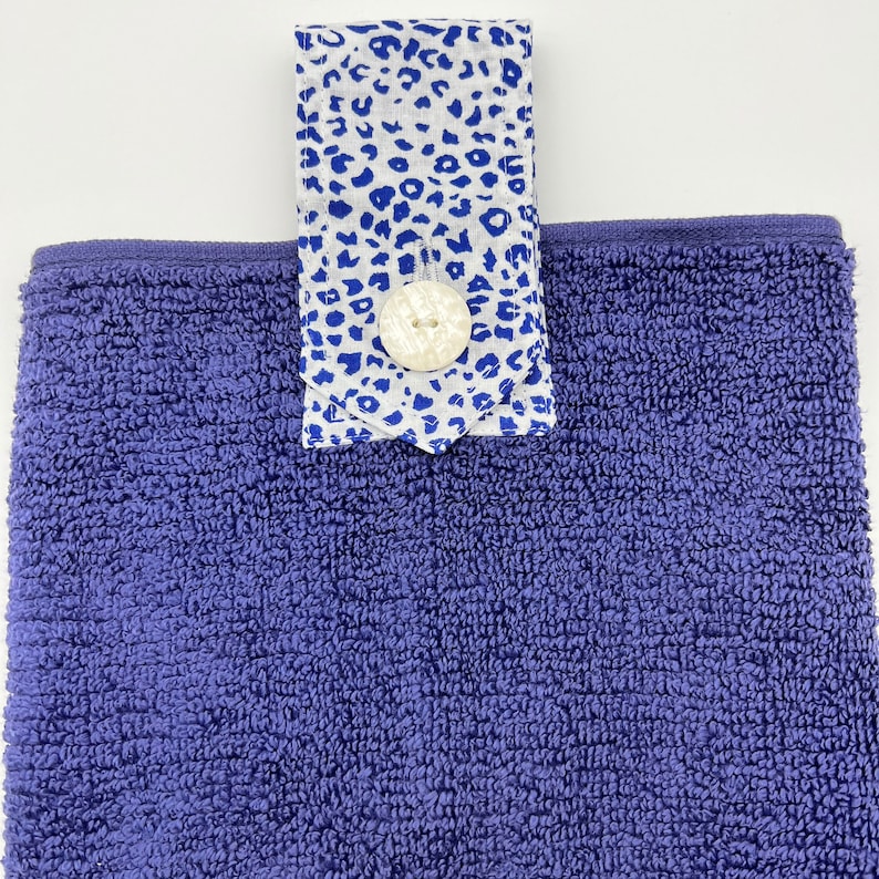 May include: A purple terry cloth hand towel with a white and blue patterned fabric loop and a white button.