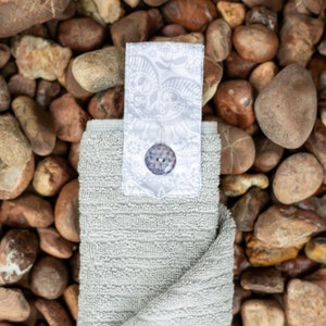 May include: A gray hand towel with a white fabric loop and a decorative button. The loop has a floral pattern. The towel is on a background of brown rocks.