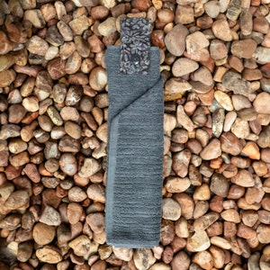 May include: A gray kitchen towel with a floral patterned fabric loop hanging from the top. The towel is folded and lying on a bed of small brown rocks.