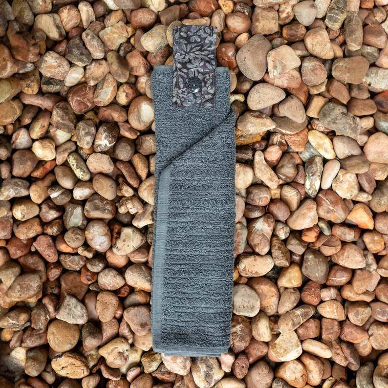 May include: A gray kitchen towel with a floral patterned fabric loop hanging from the top. The towel is folded and lying on a bed of small brown rocks.