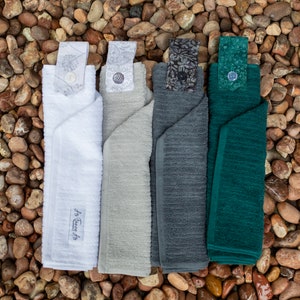 May include: Four kitchen towels with different colors and patterns. The towels are folded and have a loop on the top. The towels are on a background of small brown rocks.