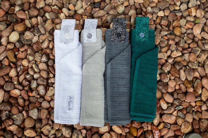 May include: Four kitchen towels with different colors and patterns. The towels are folded and have a loop on the top. The towels are on a background of small brown rocks.