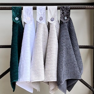 May include: Five kitchen towels hanging on a rack. The towels are in various colors and patterns, including green, white, beige, gray, and a floral pattern.