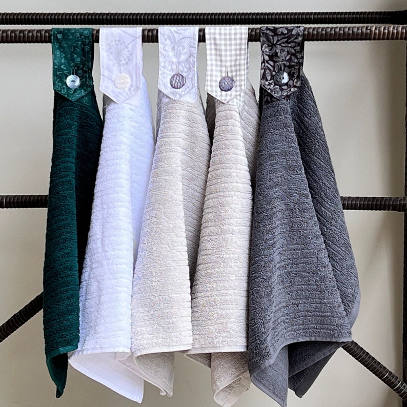 May include: Five kitchen towels hanging on a rack. The towels are in various colors and patterns, including green, white, beige, gray, and a floral pattern.
