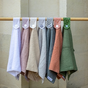 May include: Eight different kitchen towels hanging on a wooden rod. The towels are made of different fabrics and have various patterns and colours, including white, pink, beige, grey, and green.
