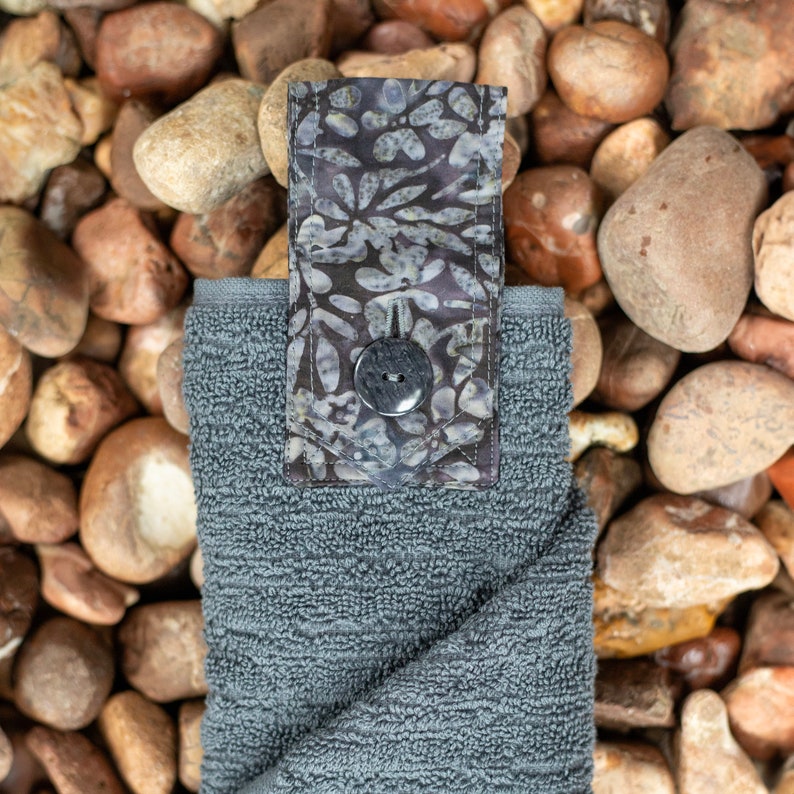 May include: A gray hand towel with a patterned fabric loop. The loop has a dark gray button.