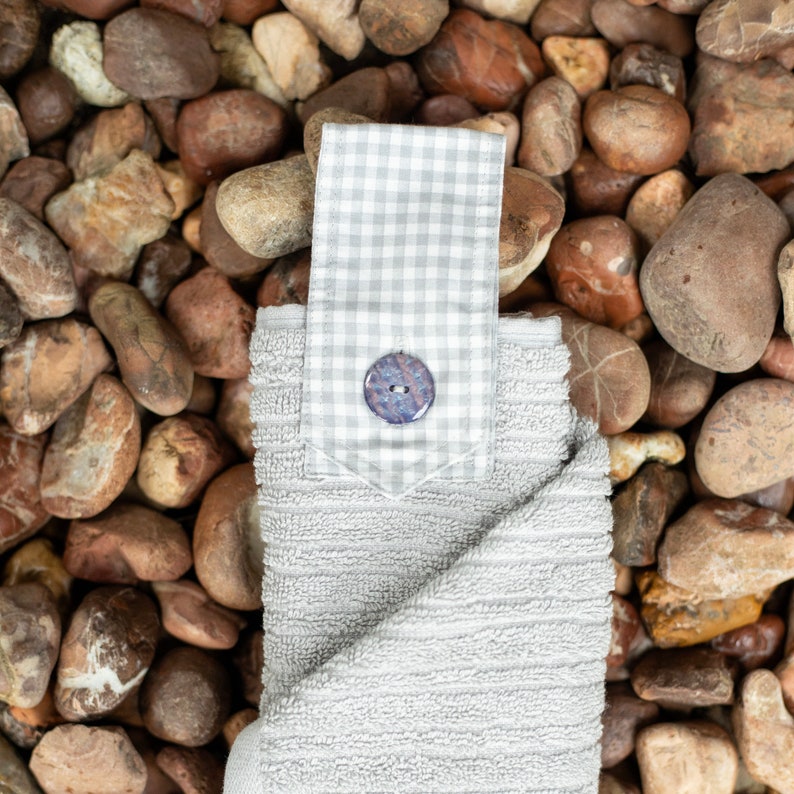 May include: A gray kitchen towel with a gingham patch and a blue button. The towel is folded and has a textured surface.
