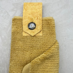 May include: A yellow kitchen towel with a decorative fabric loop and a large, round, black button. The loop has a swirl pattern.