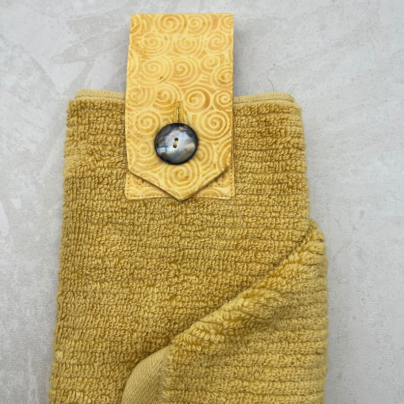 May include: A yellow kitchen towel with a decorative fabric loop and a large, round, black button. The loop has a swirl pattern.