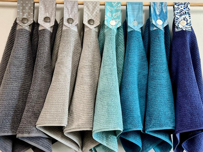 May include: Eight kitchen towels hanging on a rack. The towels are in various shades of gray and blue. The towels have different patterns on the back.