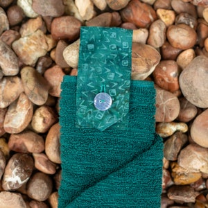 May include: A dark green hand towel with a patterned fabric loop and a white button. The towel is folded and sitting on a bed of brown and green rocks.