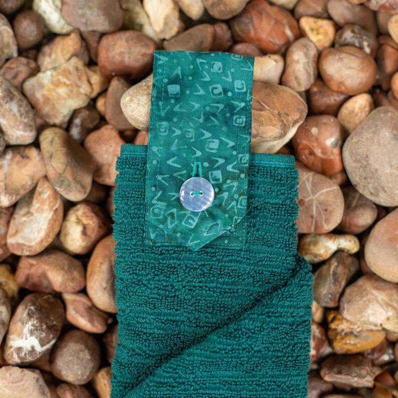 May include: A dark green hand towel with a patterned fabric loop and a white button. The towel is folded and sitting on a bed of brown and green rocks.