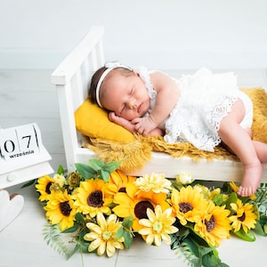May include: A newborn baby sleeps peacefully in a miniature white bed with a yellow pillow and blanket. The baby wears a white lace outfit and headband. A chalkboard displays the baby's weight as 3550 g and height as 57 cm. Sunflowers and greenery decorate the scene.