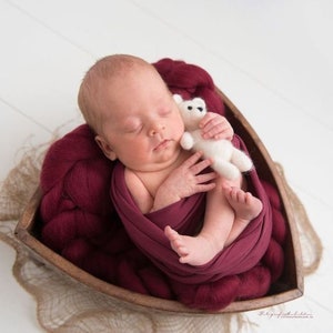 Könnte beinhalten: Ein schlafendes Neugeborenes liegt in einer Holzschale, eingehüllt in eine tiefrote Decke. Das Baby hält einen kleinen weißen Teddybären. Die Schale ruht auf einem stoffähnlichen Untergrund.