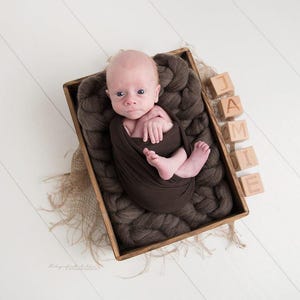 Könnte beinhalten: Ein Neugeborenes, eingewickelt in eine braune Decke, liegt in einer Holzkiste mit brauner Wolle. Holzblöcke mit der Aufschrift "JAMIE" sind neben der Kiste angeordnet. Das Baby liegt auf einer weißen Holzoberfläche.