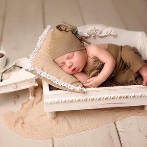 May include: A sleeping baby wearing a brown hat and outfit, resting on a small white bed with a decorative headboard. A small side table holds a cup of coffee and eyeglasses. The scene is set on a light-colored wooden floor.
