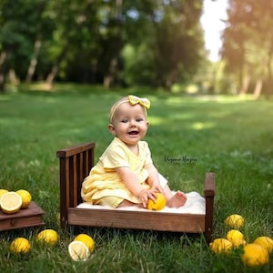 May include: A smiling baby wearing a yellow dress and headband sits on a small wooden bed in a grassy field. The baby holds a lemon, with several more lemons scattered around the bed and a small wooden box.