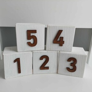 May include: Five white wooden cubes arranged in two rows, each featuring a dark brown wooden number attached to the front. The top row displays the numbers 5 and 4, while the bottom row displays the numbers 1, 2, and 3. The wood has a visible grain.