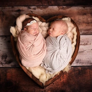 May include: Two newborn babies swaddled in pink and gray blankets, resting in a heart-shaped wooden basket filled with soft, white material. One baby wears a floral headband. The background is a rustic wooden surface.