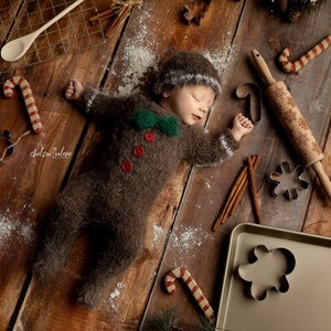 May include: A sleeping infant wearing a brown fuzzy long-sleeve romper with a green bow tie, three red buttons, and a matching hat. The outfit is surrounded by baking supplies including a rolling pin, cookie cutters, candy canes, and cinnamon sticks.