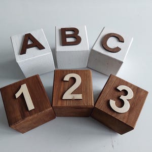 Puede incluir: Bloques de madera con letras y números. Tres bloques blancos presentan las letras A, B y C en marrón oscuro. Dos bloques marrones muestran los números 1, 2 y 3 en un tono de madera más claro. Juguete educativo.