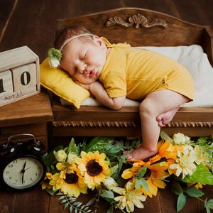 May include: A sleeping baby wearing a yellow onesie and headband rests on a small wooden bed with a yellow pillow. A wooden block calendar shows April 10. Sunflowers and greenery surround the bed, with a black alarm clock nearby.