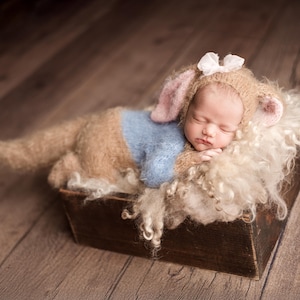 May include: A fuzzy tan and light blue knitted outfit featuring a hood with long ears and a white bow, worn by a sleeping infant. The outfit includes a textured tan body with a light blue center panel and a matching hood with pink-lined ears.