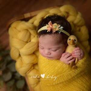 Könnte beinhalten: Ein neugeborenes Baby, eingehüllt in eine weiche, gelbe Decke, in einem Holzkörbchen. Das Baby trägt ein Blumenstirnband und hält ein kleines, gelbes Entenspielzeug. Das Bild hat eine warme, einladende Ästhetik, perfekt für Babyfotografie.