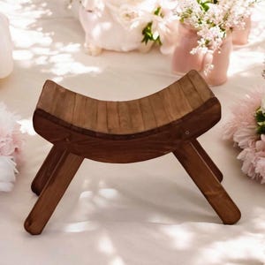 May include: A small, handcrafted wooden stool with a curved seat and angled legs. The stool is made of dark brown wood and has a natural wood grain pattern. The design is simple and elegant, suitable for various uses.
