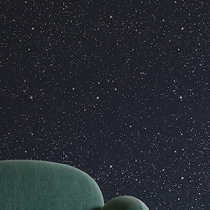 Pode incluir: Uma poltrona verde com pernas douradas fica em frente a uma parede com um papel de parede azul escuro estrelado. Uma mesa auxiliar branca com uma xícara e um pires e um relógio fica ao lado da poltrona.