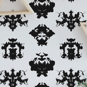 May include: A white wall with a repeating black ink blot pattern. Triangular wooden shelves are mounted on the wall. A wooden desk with a chair, a potted plant, and a camera are in front of the wall.