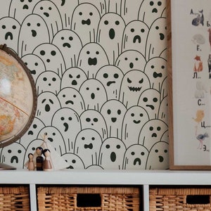 May include: A white toy house with a brown roof, a child is playing with the house. The house is in front of a white shelf with wicker baskets. The wall behind the shelf is covered in a green and white wallpaper with a pattern of ghosts.