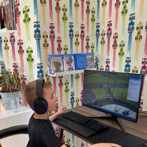 Può includere: Una persona con le cuffie gioca a un videogioco su un computer. La scrivania ha una tastiera, un mouse e un tappetino per il mouse. La stanza ha una carta da parati con auto da corsa. La persona è seduta su una sedia da gioco.