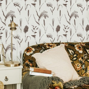 May include: A green velvet sofa with floral upholstery and a light pink throw pillow. The sofa is in front of a white wall with a floral pattern. A gold lamp with a white shade is on a white side table.