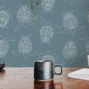 Op de afbeelding: Een zwarte keramische mok staat op een houten tafel voor een blauwe muur met een wit patroon van gestileerde zeeschildpadden.
