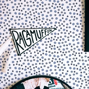 May include: A white pennant with the words "Ragamuffin" written in black marker on a white background. The pennant is hanging on a wall with a blue and grey polka dot pattern.