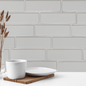 Op de afbeelding: Een witte keramische mok met een witte keramische schotel op een houten oppervlak voor een grijze betegelde muur. Een potplant en een vaas met gedroogde grassen staan op het oppervlak.