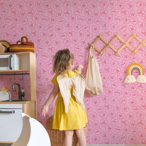 May include: A pink wallpaper with a repeating pattern of red hearts with different sayings inside. The sayings include "Kiss Me", "Marry Me", "True Love", "Sweet", "Call Me", "One Love", and "You and Me". A young girl is standing in front of the wallpaper, wearing a yellow dress and angel wings.