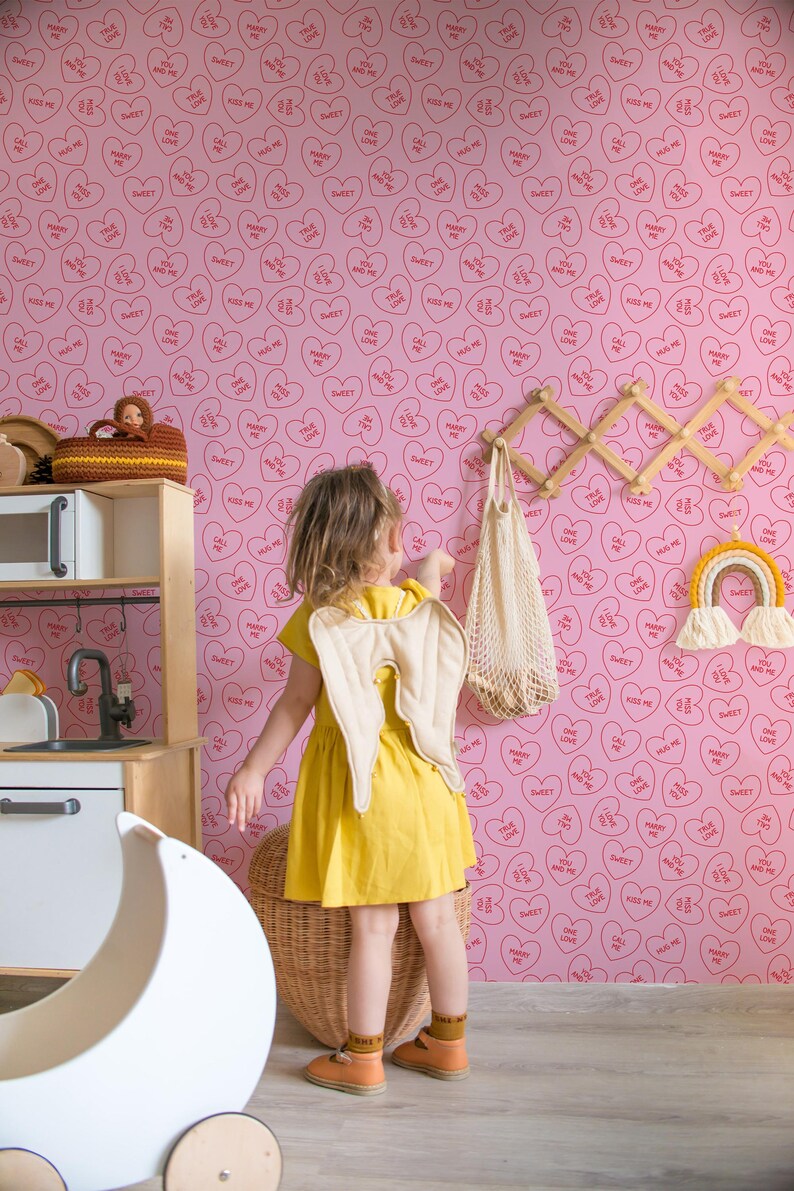 May include: A pink wallpaper with a repeating pattern of red hearts with different sayings inside. The sayings include "Kiss Me", "Marry Me", "True Love", "Sweet", "Call Me", "One Love", and "You and Me". A young girl is standing in front of the wallpaper, wearing a yellow dress and angel wings.
