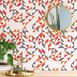 Può includere: Un muro bianco con una carta da parati a motivi geometrici con triangoli arancioni, rossi e neri. Uno specchio dorato con una cornice decorativa è appeso al muro. Una pianta in vaso, una pila di libri e un vassoio dorato con oggetti decorativi sono su un tavolo di legno davanti al muro.