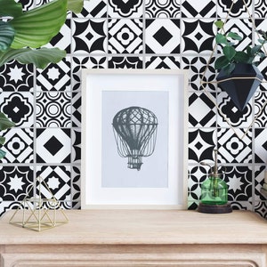 Puede incluir: Papel tapiz de azulejos geométricos en blanco y negro con una impresión enmarcada de un globo aerostático, una escultura geométrica de metal dorado, una botella de spray de vidrio verde, una maceta colgante geométrica negra y dorada con una planta verde y una pila de libros sobre una mesa de madera.