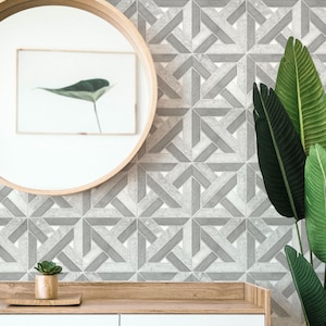 Op de afbeelding: Een witte commode met twee laden en een houten blad. De commode staat voor een muur met een geometrisch patroon van grijze en witte tegels. Een kleine plant in een pot staat op de commode. Een ronde spiegel met een houten lijst hangt aan de muur boven de commode. Een grote groene plant staat aan de rechterkant van de afbeelding.