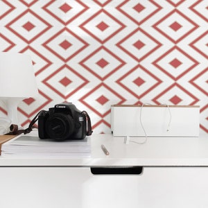 Op de afbeelding: Een wit bureau met een zwarte camera, een witte kabelorganizer en een potlood erop. De muur achter het bureau heeft een rood en wit geometrisch patroon.
