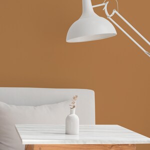 Pode incluir: Uma luminária de mesa branca com uma grande cúpula arredondada está posicionada acima de uma pequena mesa branca. Um pequeno vaso branco com flores secas está sobre a mesa. O fundo é de cor marrom quente.